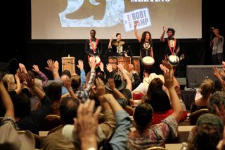 An audience raises their hands, mimicking the motion of the drummers on stage.