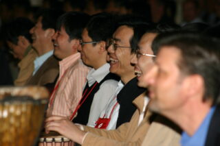 A group of people in red lanyards smile as they play the drums.