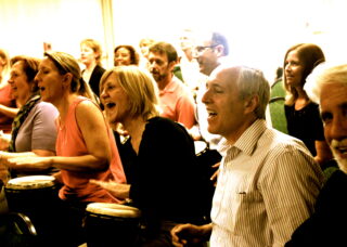 A group of people lean to the side while playing hand drums in front of them.